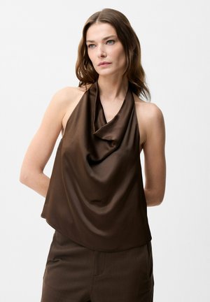 Vrouw met lang bruin haar die een mouwloze donkerbruine satijnen haltertop en bijpassende broek draagt tegen een neutrale achtergrond.
