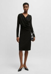 Black wrap dress with long sleeves, knee-length, featuring a gathered side detail. Paired with black heeled shoes and a small handbag.