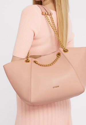 Kvinde i en lyserød ribstrikket kjole bærende en stor blush-lyserød Guess-håndtaske med gyldne kædehåndtag over skulderen.