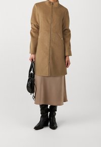 Cappotto in lana beige con colletto alto e zip frontale, abbinato a una gonna satin beige di media lunghezza e stivali neri al ginocchio. Borsa nera.