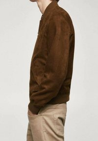 Chaqueta bomber de ante marrón con cuello y puños acanalados, que presenta bolsillos laterales y cierre de cremallera, mostrada de lado.