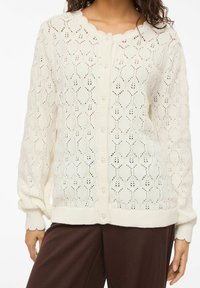 Femme portant un cardigan blanc en maille avec un motif losange et des boutons en perle, associé à un pantalon marron ample.