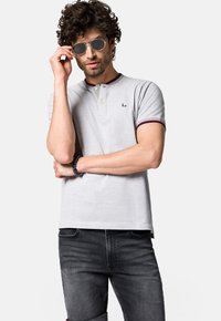 Szara koszulka polo z kołnierzykiem na dwa guziki, prążkowanymi czerwonymi i niebieskimi akcentami oraz detalem z logo; w połączeniu z ciemnymi dżinsami i okularami przeciwsłonecznymi.