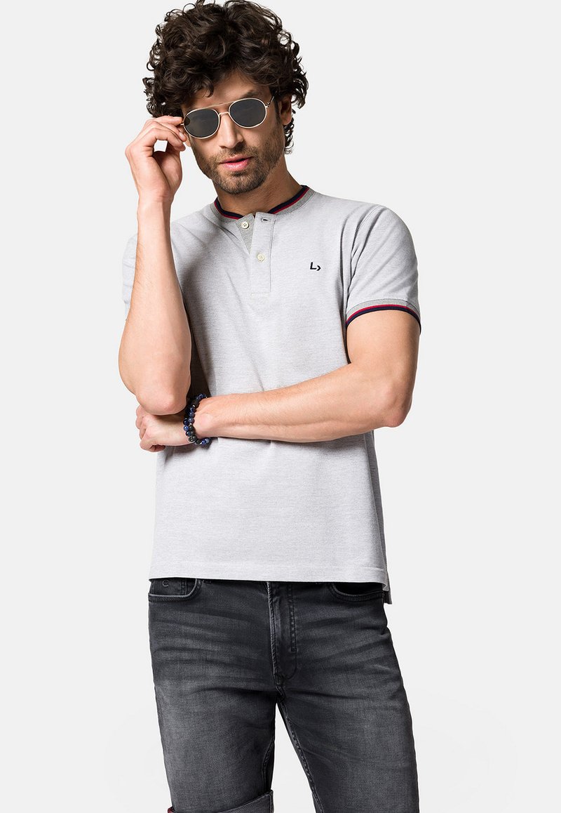 Szara koszulka polo z kołnierzykiem na dwa guziki, prążkowanymi czerwonymi i niebieskimi akcentami oraz detalem z logo; w połączeniu z ciemnymi dżinsami i okularami przeciwsłonecznymi.