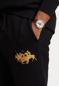 Pantaloni sportivi neri con un evidente logo ricamato dorato di un polo con tre cavalieri a cavallo. La vita è elastica e presenta tasche laterali.