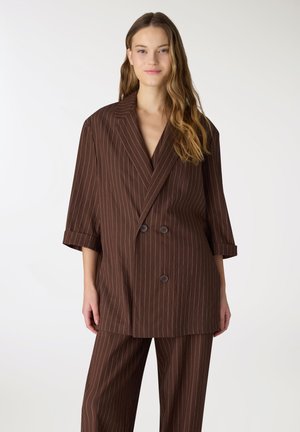 Donna con i capelli lunghi che indossa una giacca doppiopetto marrone a righe larghe e pantaloni coordinati, in piedi davanti a uno sfondo chiaro e semplice.