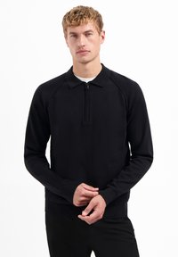Zwarte pullover met een ritssluiting bij de kraag, gemaakt van een zachte stof. Heeft lange mouwen en een aangesloten ontwerp met geribbelde accenten bij de manchetten en de zoom.