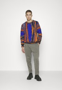 Bunter Strickpullover mit kräftigen Mustern in Orange, Blau und Schwarz, kombiniert mit hellgrauen Hosen und schwarzen klobigen Schuhen.
