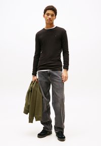 Schwarzes Langarmhemd mit weißem Kragen, graue Jeans, hält einen grünen Hoodie. Schwarze Sneakers mit strukturiertem Detail, vor neutralem Hintergrund stehend.