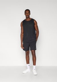 Μαύρο αθλητικό τοπ χωρίς μανίκια με υφή δίχτυ, συνδυασμένο με μαύρα σορτς που φέρουν λεπτομέρεια λογότυπου, φορεμένο με λευκές κάλτσες και αθλητικά παπούτσια.