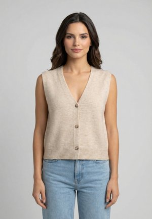 Mujer con cabello largo y oscuro, vistiendo un chaleco de punto sin mangas color beige abotonado y jeans de tiro alto azul claro, de pie contra un fondo liso.