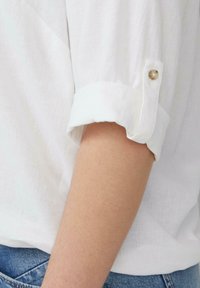 Witte linnen blouse met opgerolde mouwen, voorzien van een knoopaccent op de manchette. De stof lijkt licht en heeft een lichte textuur.