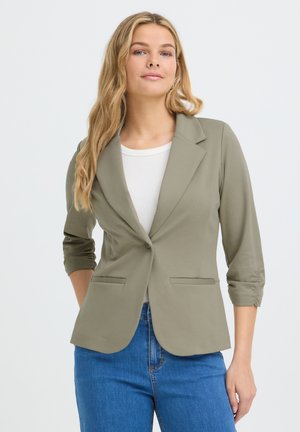 Femme aux longs cheveux blonds portant un blazer vert olive clair par-dessus un top blanc et un jean bleu, debout avec une main dans une poche.