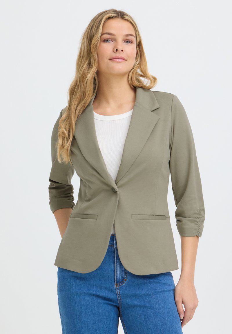 Femme aux longs cheveux blonds portant un blazer vert olive clair par-dessus un top blanc et un jean bleu, debout avec une main dans une poche.