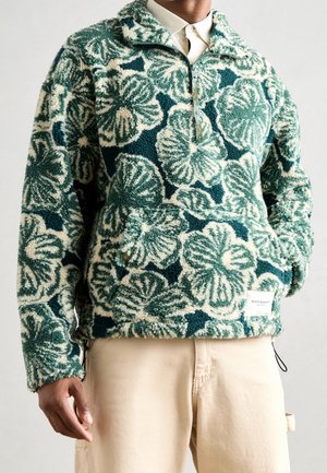 Personne portant un pull polaire texturé vert et blanc à motifs floraux avec une fermeture éclair partielle et un pantalon beige, mains détendues le long du corps.