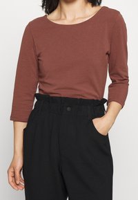 Rostfärgad, åtsittande topp med en bred rund halsringning och trekvartsärmar, tillsammans med högmidjade svarta byxor med fickor.