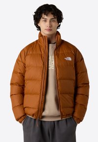 Brun oppustet jakke med høj krave og lynlås. Har et quiltet mønster og et lille hvidt logo på brystet. Bæret over en beige sweater.
