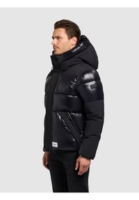 Črna puhovka s sijočim zaključkom, žepki z zadrgo in velikim kapuco. Ima vodoravne šive in znamkasto etiketo na spodnjem delu.