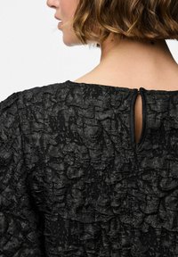 Svart texturerad klänning med en skrynklig yta. Rund halsringning i ryggen med en liten nyckelhålsöppning och en diskret dragkedja.
