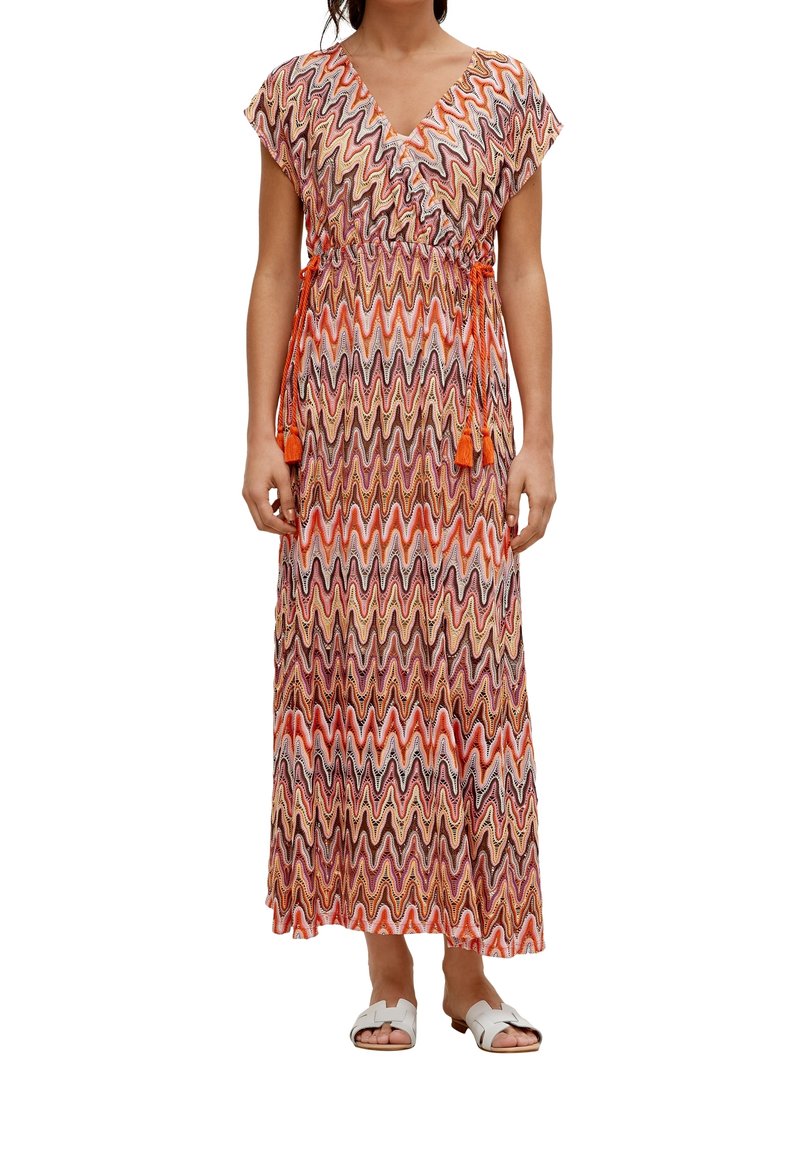 Robe maxi orange, rouge et marron à motif zigzag ; présente un décolleté en V, des manches courtes et une taille nouée avec des franges. Assortie avec des sandales blanches.