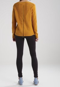 Senfgelber Strickpullover mit strukturiertem Zickzackmuster, Reißverschlussteil im Rücken, gerippter Saum. Kombiniert mit schwarzen, enganliegenden Hosen und hellblauen Absätzen.