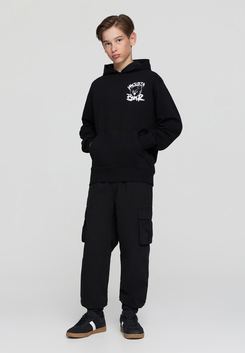 Felpa nera con tasca a marsupio frontale e logo grafico bianco. Abbinata a pantaloni cargo neri con tasche laterali. Sneakers nere con strisce bianche.