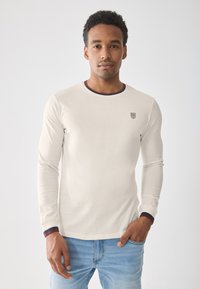 Camisa de manga larga color beige claro con un cuello y puños en contraste de color navy, que incluye un pequeño logo en el lado izquierdo del pecho. Se lleva con jeans de mezclilla azul.