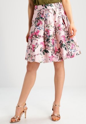 Femme portant une jupe fleurie rose mi-longue et des sandales à talons hauts métalliques couleur or rose, debout devant un fond uni.