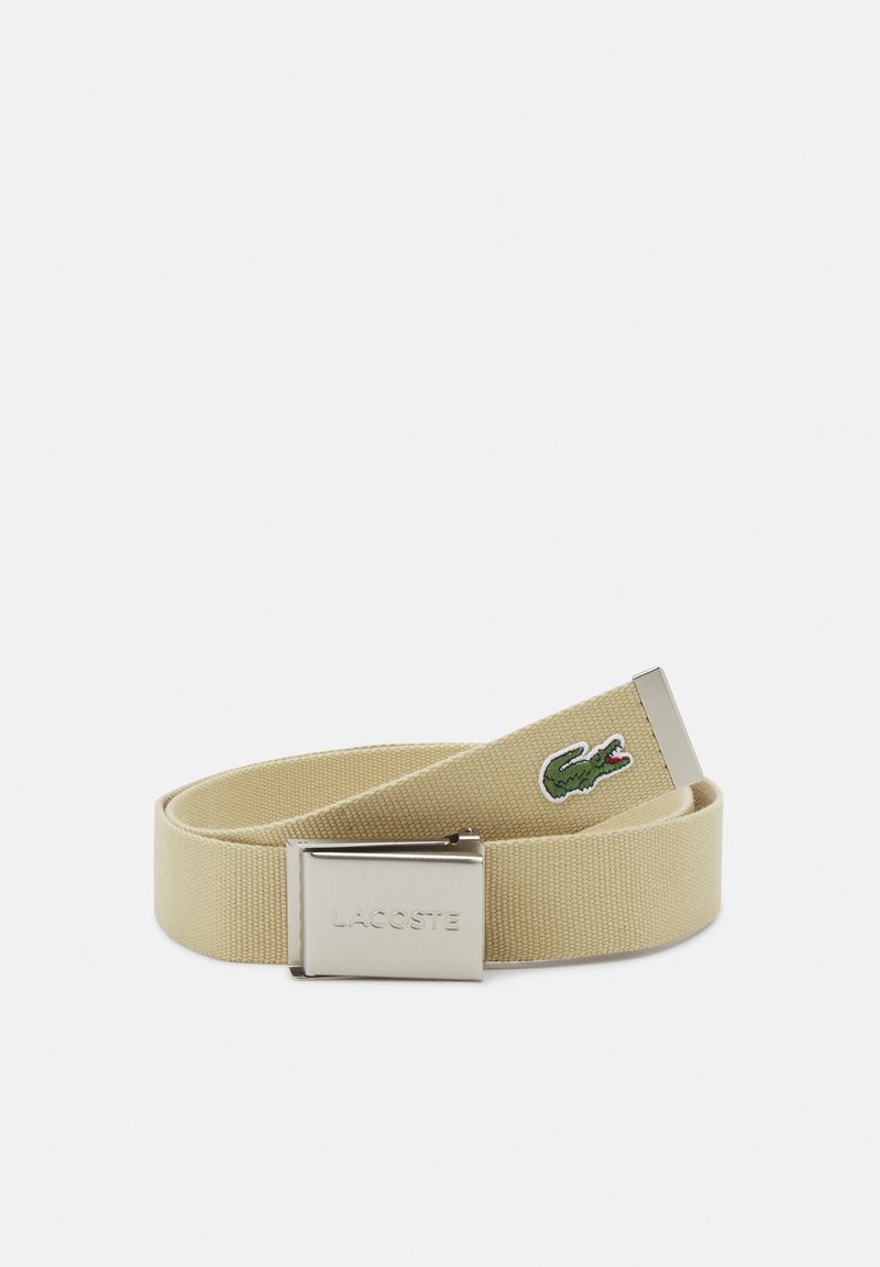 Béžový pletený opasok s kovovým sponou, ktorý má vyrazené logo "LACOSTE" a detail vyšívaného zeleného krokodíla. Hladký, textúrovaný povrch.