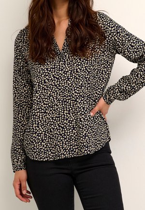 Femme aux longs cheveux bruns portant un chemisier noir avec de petits motifs floraux blancs et un pantalon noir, une main sur la hanche, sur un fond clair.