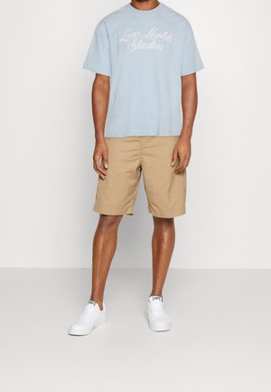 Ljusblå bomulls-t-shirt med texten "Love Lights Studios" och beige chinoshorts. Modellen bär vita sneakers. Enkelt och avslappnat outfit.