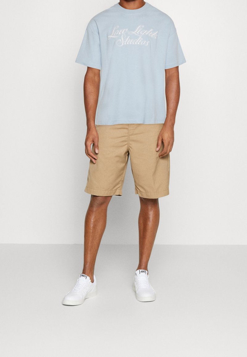 T-shirt en coton bleu clair avec le texte "Love Lights Studios" et shorts chino beiges. Le modèle porte des baskets blanches. Tenue simple et décontractée.