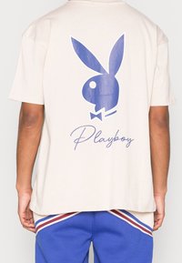 Person som bär en beige t-shirt med en stor blå Playboy-kaninlogo och kursiv "Playboy"-text på ryggen, i kombination med blå och randiga byxor.