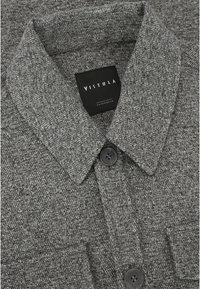 Szary kołnierz w wzór w kieszeń herringbone z czarnym guzikiem i etykietą "VISTULA OUTERWEAR". Tkanina ma teksturalny splot.