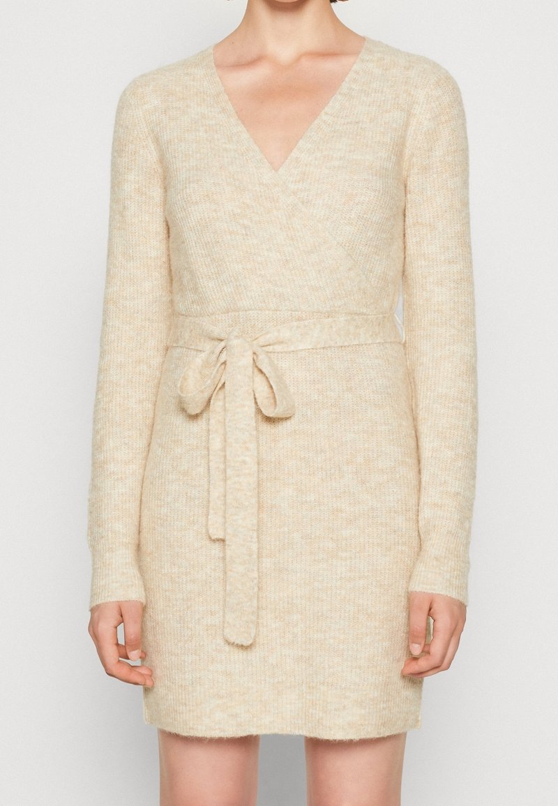 Robe cache-cœur tricotée beige avec manches longues, col en V et ceinture nouée à la taille. Texture douce avec un motif chiné subtil.