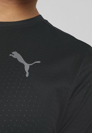 Primer plano de una camiseta deportiva negra con pequeñas perforaciones y un logo gris de Puma en la parte superior del pecho, cerca del hombro.