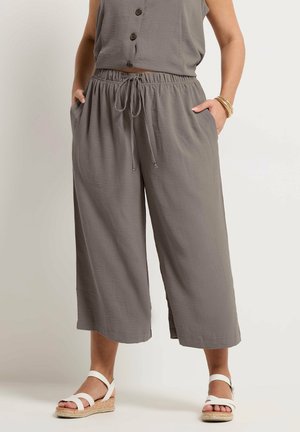 Femme portant un pantalon ample gris coupé au-dessus de la cheville avec une taille élastique à cordon, associé à des sandales blanches à brides et un haut gris boutonné.