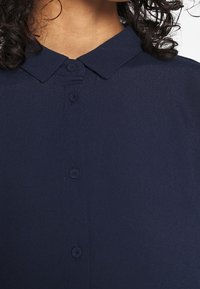 Femme portant une chemise boutonnée bleu foncé avec un col pointu et des cheveux bouclés foncés visibles au niveau du cou.