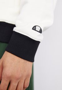 Witte sweatshirt met zwarte geribbelde manchetten. Bevat een klein zwart logo op de manchette. Glad textuur en casual ontwerp.