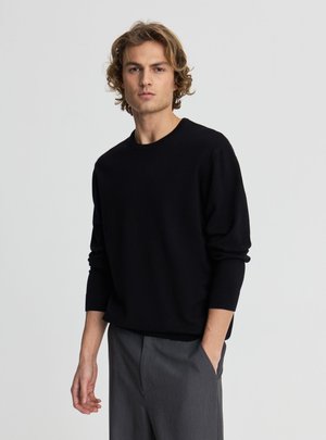 Giovane uomo con capelli biondi ondulati che indossa un maglione nero a maniche lunghe e pantaloni grigi, in piedi con una mano in tasca davanti a uno sfondo grigio uniforme.