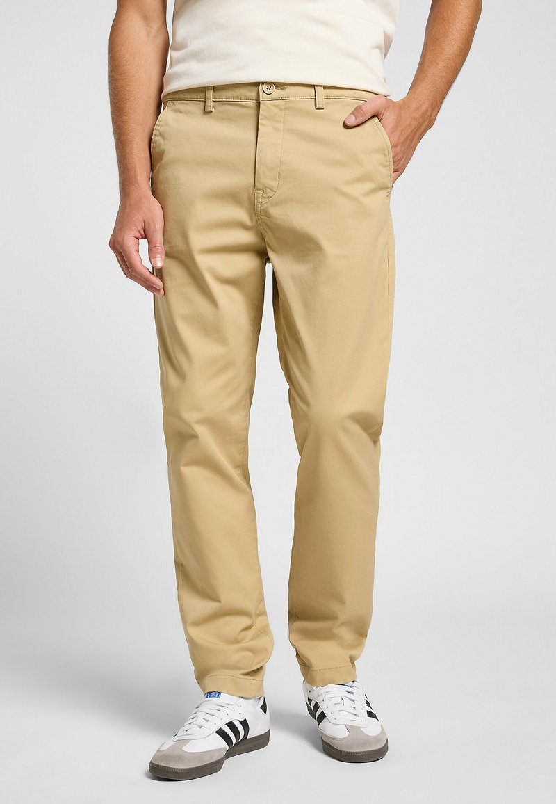 Chinos beige fabricados en algodón, con un corte recto, bolsillos frontales y cierre de botón. Llevados con zapatillas deportivas blancas y grises.