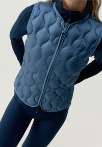 Chaleco acolchado azul marino con cuello alto y cremallera frontale, que presenta un tejido suave y texturizado con un diseño de patrón ondulado. Se lleva sobre leggings a juego.