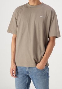 Brązowy bawełniany T-shirt z krótkimi rękawami, okrągłym dekoltem i subtelnym logo "Hollister" na piersi. Charakteryzuje się luźnym krojem i miękką fakturą.