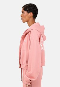 Felpa rosa cropped con zip frontale, dettagli con cordino e testo bianco sulla manica sinistra. Tessuto morbido e liscio con una vestibilità rilassata.