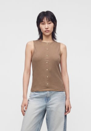 Persona con capelli neri lisci indossa un top marrone senza maniche con bottoni e jeans azzurri, in piedi contro uno sfondo bianco semplice.