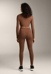 Smeđa sportska odjeća, uključujući dugi rukav i visokog struka helanke, izrađena od rastezljivog materijala. Nošena s crnim sportskim tenisicama.