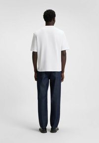 Hvid kortærmet t-shirt med rund halsudskæring, kombineret med mørkeblå denimbukser og sorte sko. Synlige detaljerne på bagsiden inkluderer sømme.