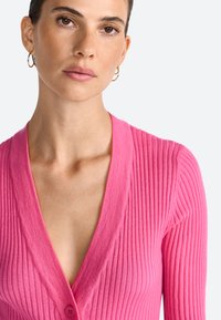 Cardigan rose à côtes avec un décolleté en V profond, doté d'une fermeture à boutons et d'un tissu texturé. Il a de longues manches et un design ajusté.