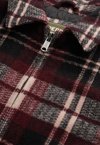 Czerwono-czarna kurtka w kratę z zamkiem błyskawicznym, wykonana z wełny i posiadająca kołnierz. Szare i beżowe akcenty w wzorze.