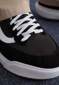 Fekete sneaker szintetikus anyagból, fehér fűzővel. Jellemzője a texturált fekete felsőrész, fehér kiemelések és a gumitalp.